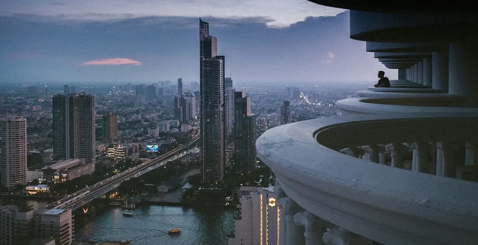 A city skyline at dusk with tall buildings and a river. A person stands on a curved balcony in the foreground, gazing at the urban landscape. The sky is a gradient of blue and purple, enhancing the citys evening lights in Bangkok, Thailand.