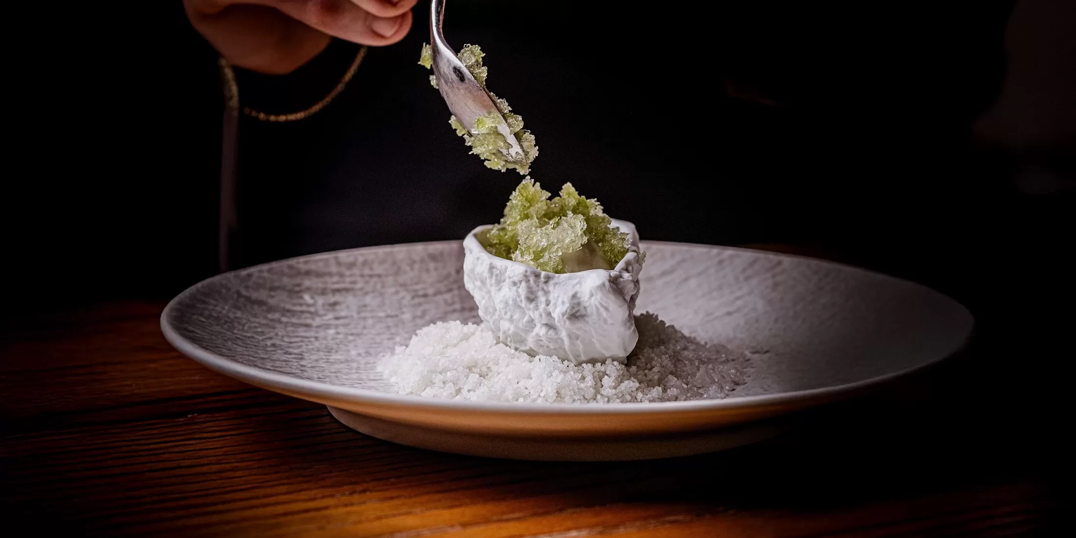 A person spoons green granita into a shell-shaped dish on a bed of ice on a plate. The scene is dimly lit, highlighting the texture of the granita and the ceramic details.
