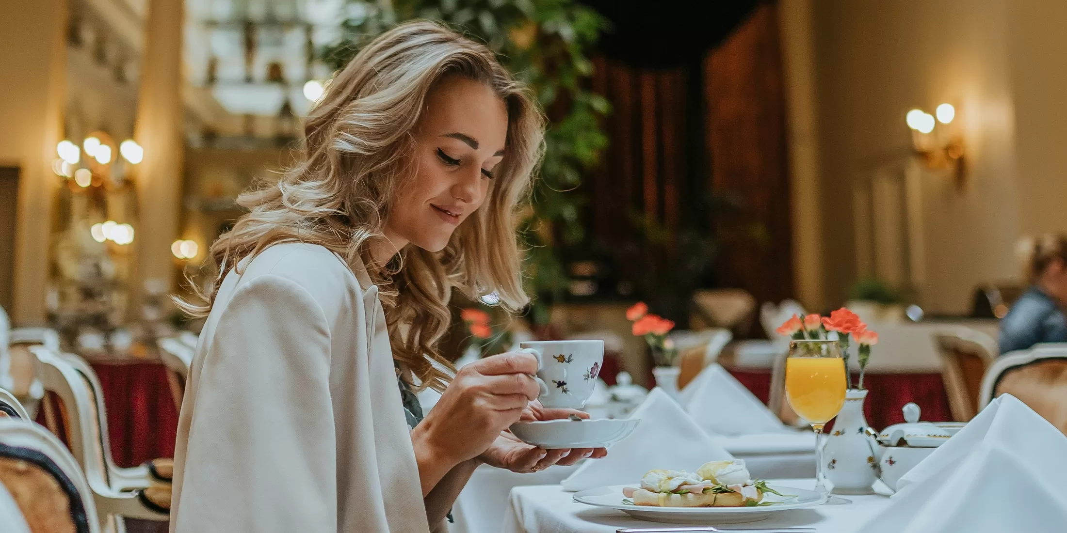 A woman with long hair is sitting at a table in a restaurant, holding a cup and saucer. She has a light jacket on and is smiling. The table has white tablecloths, set with a plate of food, a glass of orange juice, and small flowers.