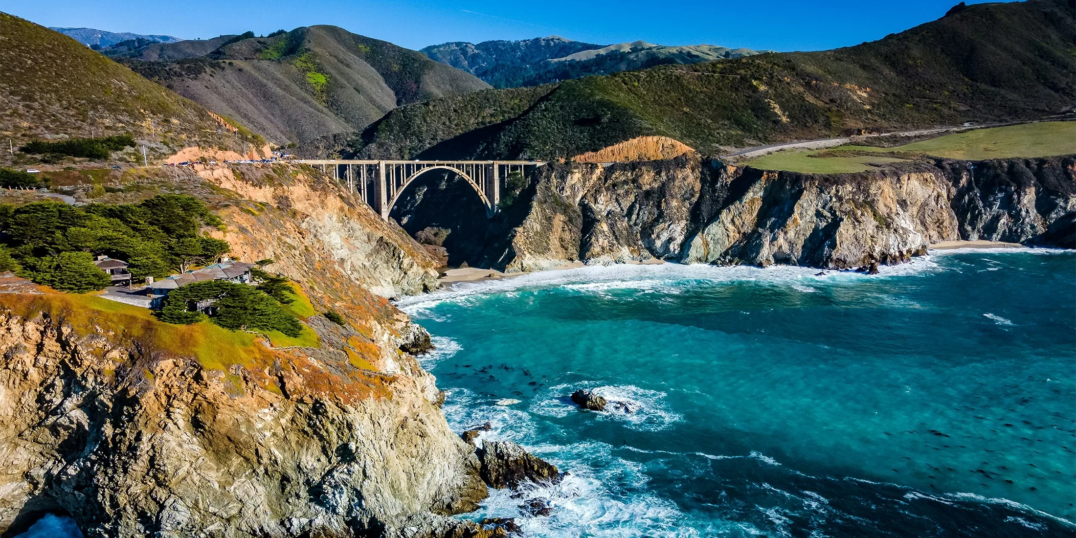 Aerial view of Bixby Creek Bridge spanning a rocky coastline along Big Sur, California, with turquoise ocean waves crashing against cliffs and green hills in the background under a clear blue sky.