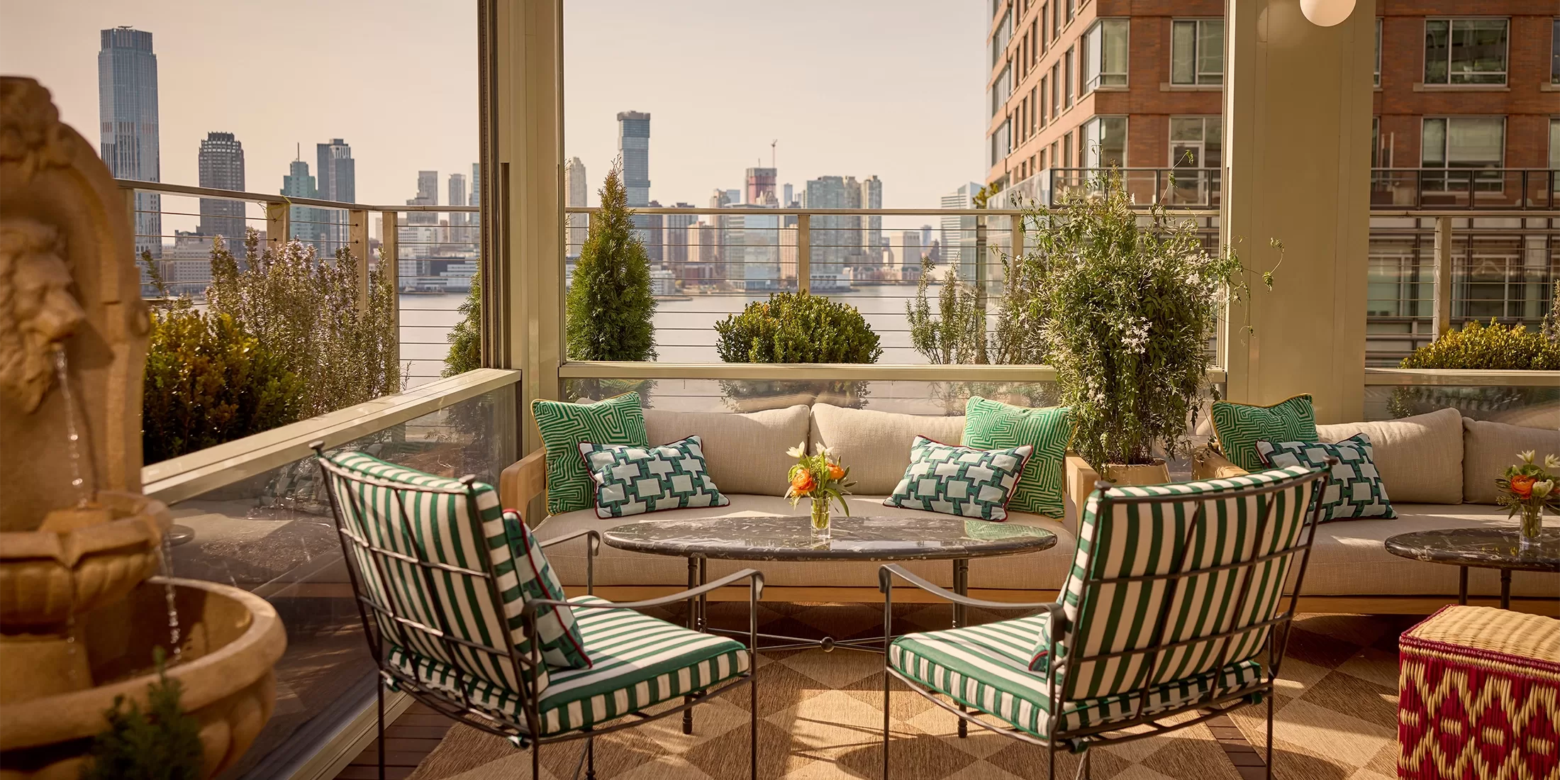 A stylish outdoor patio with striped chairs, a cushioned sofa, and a round table topped with flowers, surrounded by greenery and city buildings in the background under a clear sky.