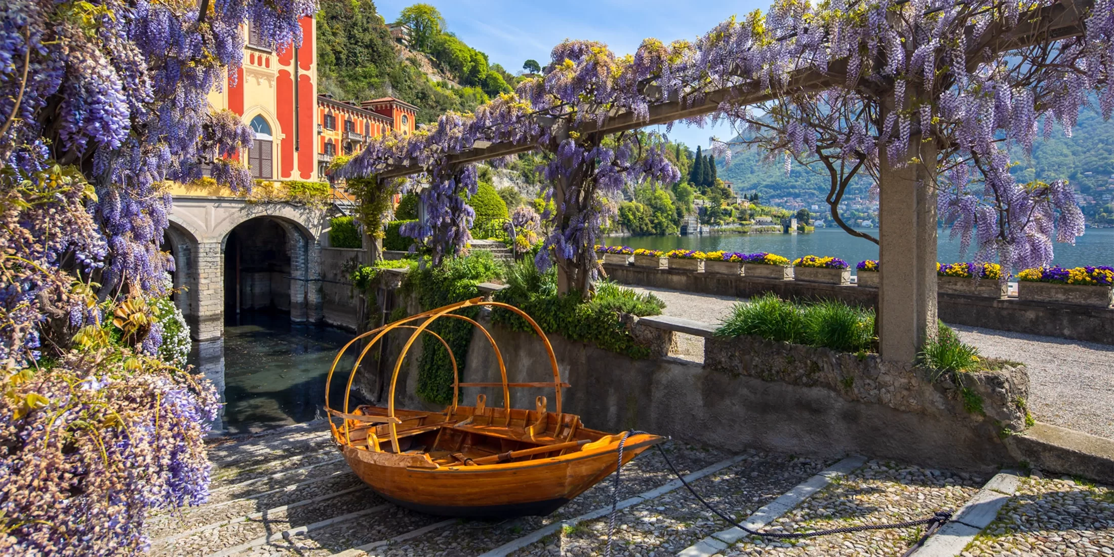 A wooden boat is moored by stone steps under a pergola covered with blooming purple wisteria, near a colorful building by a tranquil lake surrounded by lush hills and gardens.