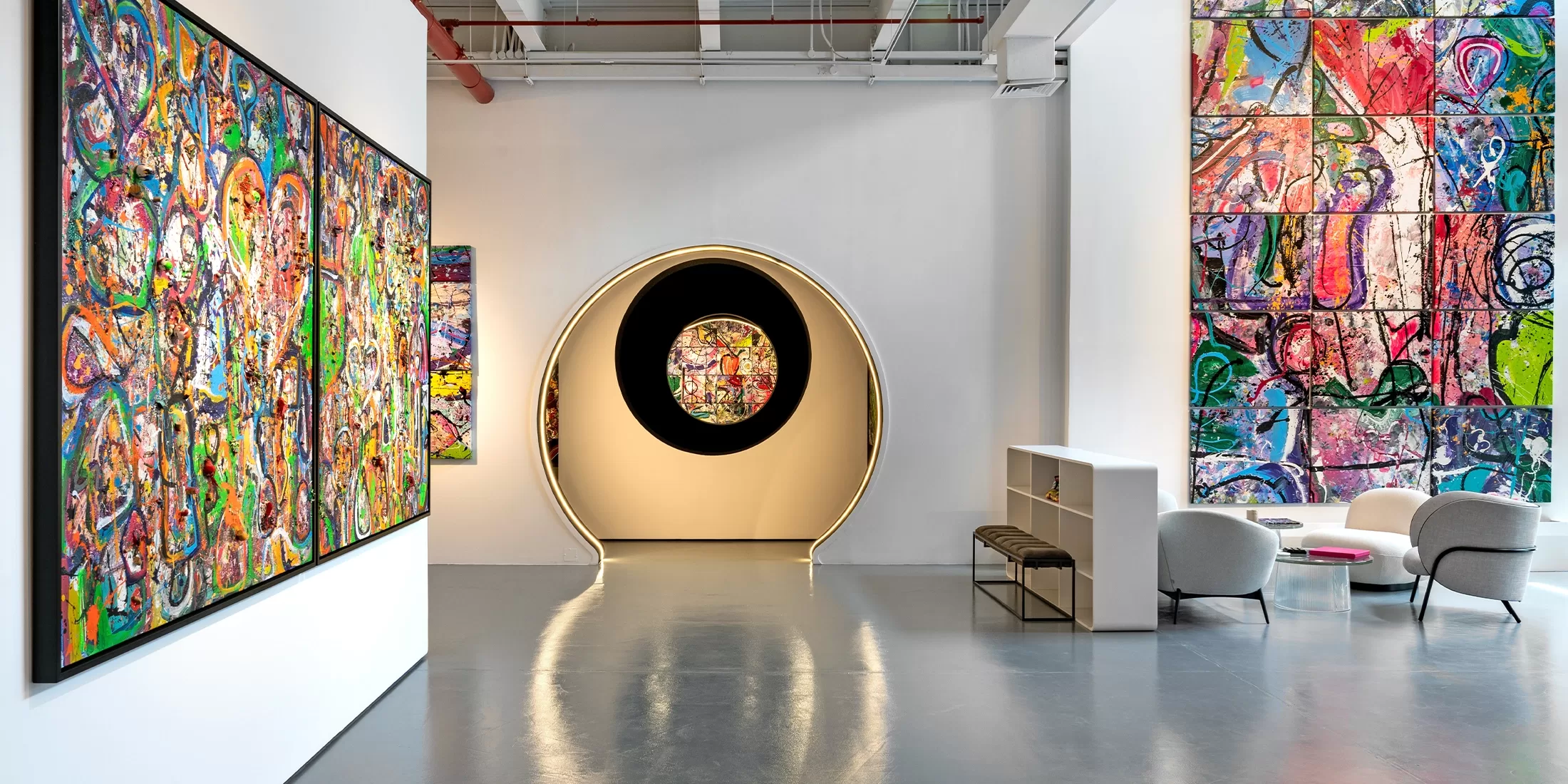 A modern art gallery with colorful abstract paintings on the walls, a circular illuminated doorway in the center, and a small seating area with chairs and a table on the right. The space features high ceilings and sleek, polished floors.