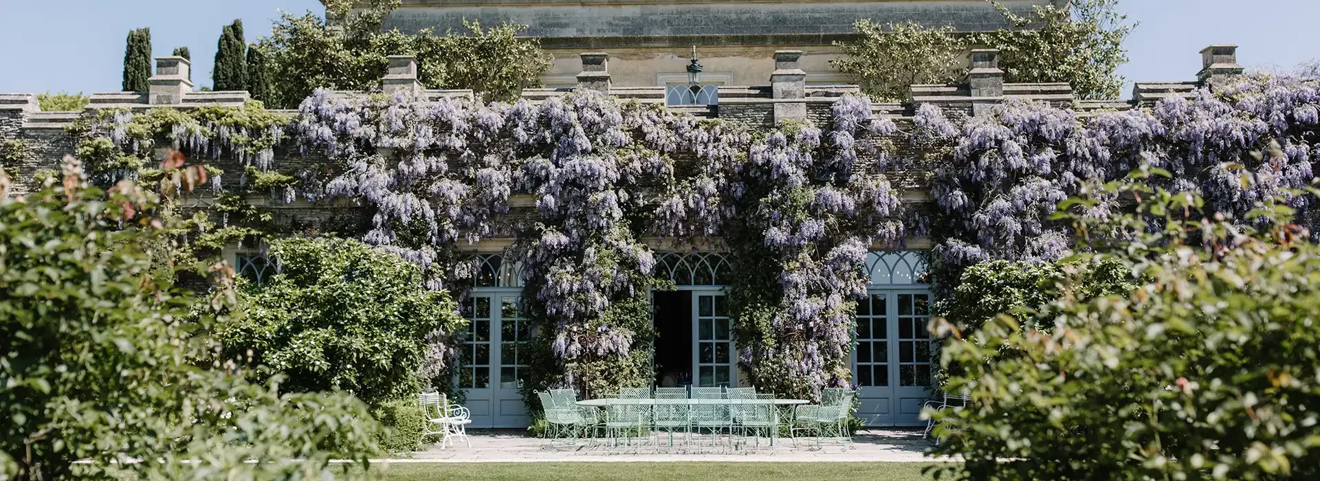 A grand, historic building with a stone facade is adorned with cascading purple wisteria plants. There are multiple arched windows and a large patio area featuring green metal furniture, surrounded by lush greenery.