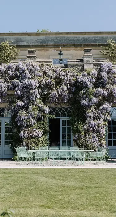 A grand, historic building with a stone facade is adorned with cascading purple wisteria plants. There are multiple arched windows and a large patio area featuring green metal furniture, surrounded by lush greenery.