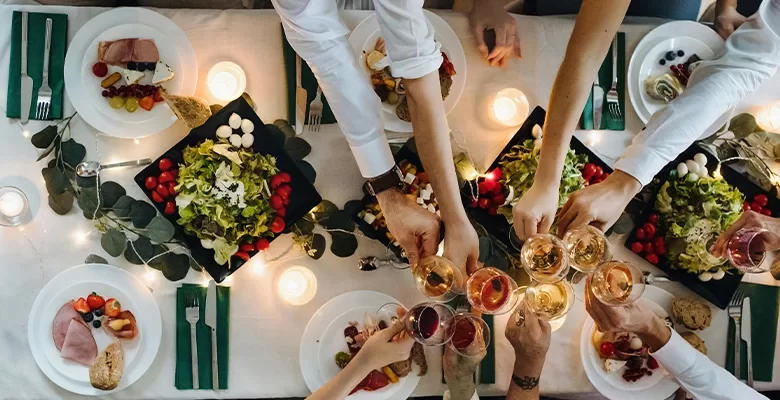 Areal view of a group of individuals hands joyfully toasting with glasses at a beautifully set dinner table filled with food.