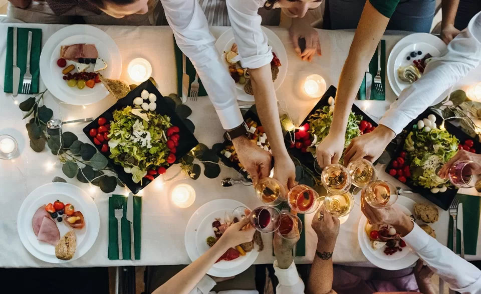 Areal view of a group of individuals hands joyfully toasting with glasses at a beautifully set dinner table filled with food.